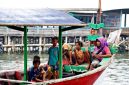 Keluarga dengan dua anak kecil sedang bermain di tepi pantai berpasir putih, menikmati momen kebersamaan di destinasi liburan tropis. Mereka terlihat bahagia dengan latar belakang laut biru jernih dan pohon kelapa yang melambai.