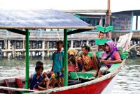 Keluarga dengan dua anak kecil sedang bermain di tepi pantai berpasir putih, menikmati momen kebersamaan di destinasi liburan tropis. Mereka terlihat bahagia dengan latar belakang laut biru jernih dan pohon kelapa yang melambai.