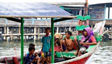 Keluarga dengan dua anak kecil sedang bermain di tepi pantai berpasir putih, menikmati momen kebersamaan di destinasi liburan tropis. Mereka terlihat bahagia dengan latar belakang laut biru jernih dan pohon kelapa yang melambai.