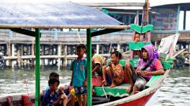 Keluarga dengan dua anak kecil sedang bermain di tepi pantai berpasir putih, menikmati momen kebersamaan di destinasi liburan tropis. Mereka terlihat bahagia dengan latar belakang laut biru jernih dan pohon kelapa yang melambai.