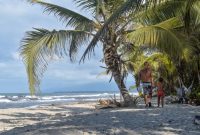 Sebuah keluarga sedang menikmati keindahan pantai berpasir putih di Sumbawa yang dihiasi dengan perbukitan hijau, menciptakan suasana liburan yang damai dan menyegarkan. Anak-anak tampak bermain air di tepi pantai, sementara orang dewasa bersantai menikmati panorama alam yang masih sangat asri.