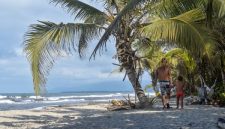 Sebuah keluarga sedang menikmati keindahan pantai berpasir putih di Sumbawa yang dihiasi dengan perbukitan hijau, menciptakan suasana liburan yang damai dan menyegarkan. Anak-anak tampak bermain air di tepi pantai, sementara orang dewasa bersantai menikmati panorama alam yang masih sangat asri.