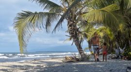 Sebuah keluarga sedang menikmati keindahan pantai berpasir putih di Sumbawa yang dihiasi dengan perbukitan hijau, menciptakan suasana liburan yang damai dan menyegarkan. Anak-anak tampak bermain air di tepi pantai, sementara orang dewasa bersantai menikmati panorama alam yang masih sangat asri.